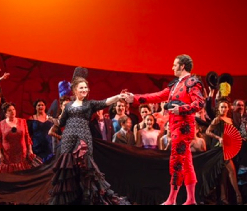 Escamillo in a red matador costume takes the hand of Carmen onstage.
